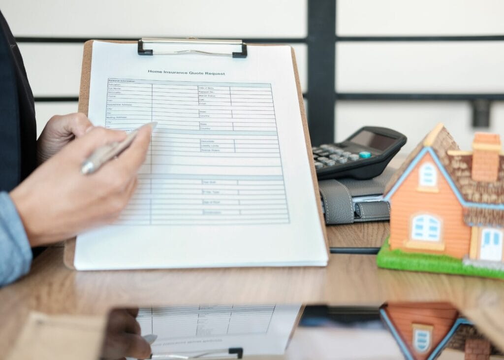 Person filling out a home insurance quote request form, next to a calculator and model house.