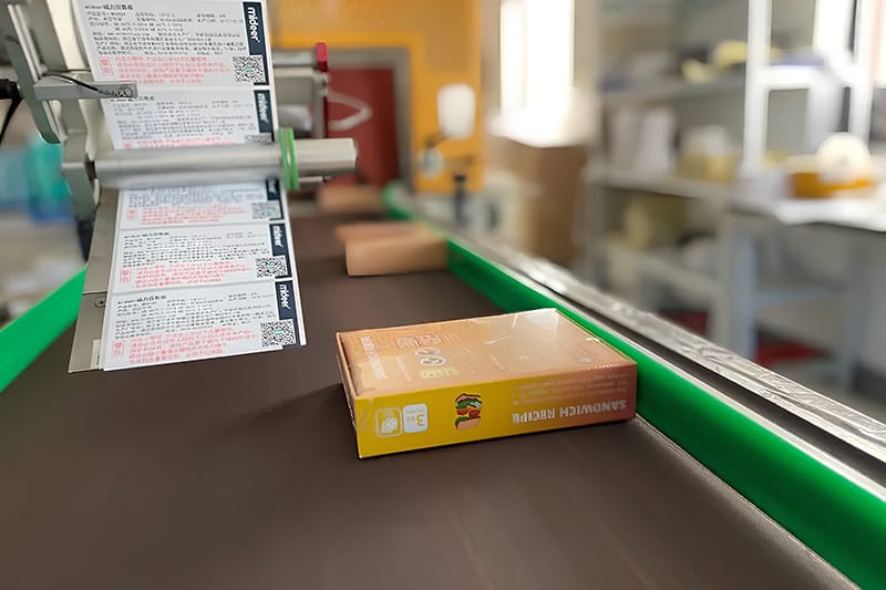 Automated labeling machine applying labels to sandwich recipe boxes on a conveyor belt.