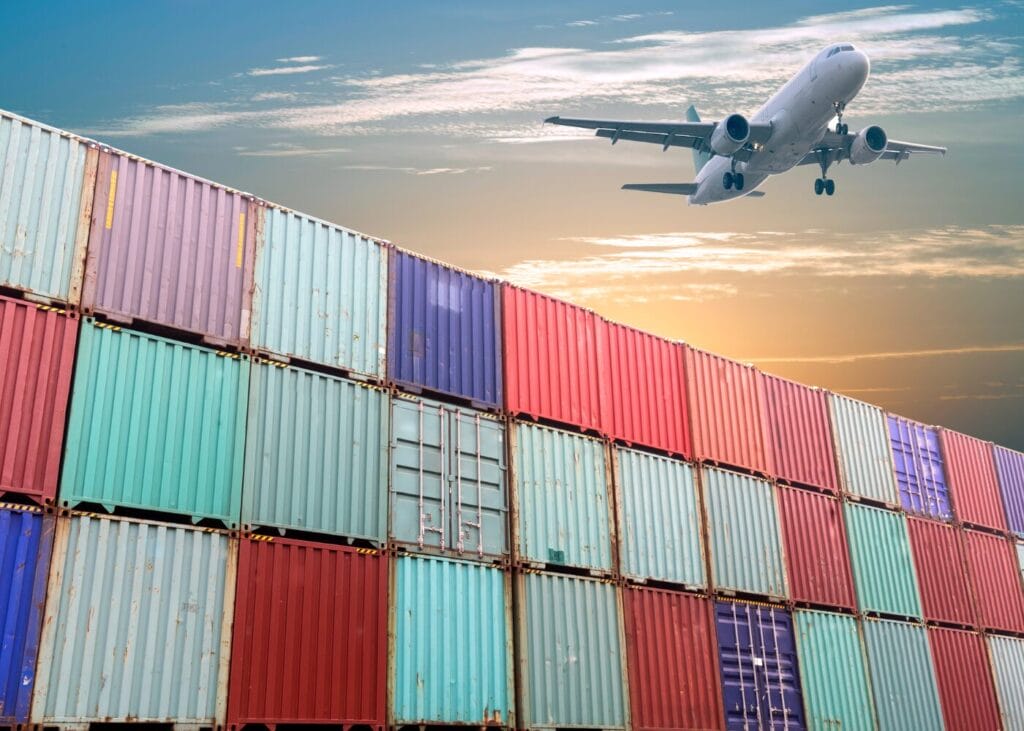 Cargo plane flying over stacked shipping containers at sunset. Global logistics and freight transport.